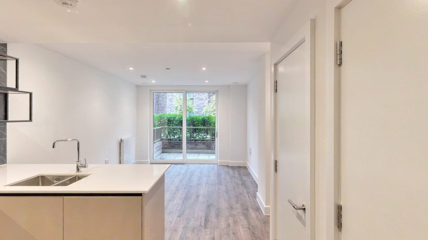 Apartment 2-1-8 from the hall: a white kitchen island with a stainless sink, oak-effect flooring running to glass doors onto the balcony and the hedges of the podium garden