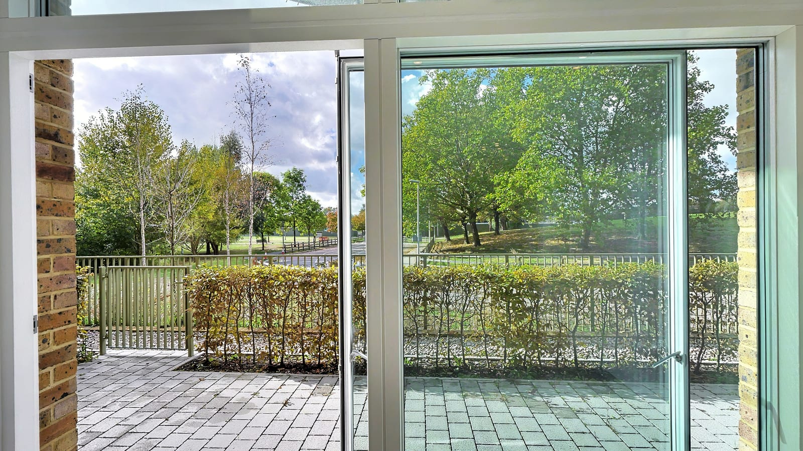Living room in apartment E1.00.03 opening through full-height glass doors onto a paved patio, a hedge and the trees of South Cator Park