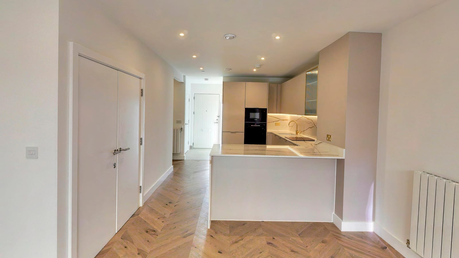 Ground floor of townhouse TH05: chevron oak flooring running the length of the open-plan room to a kitchen peninsula with a marble-effect worktop, the hall and front door beyond